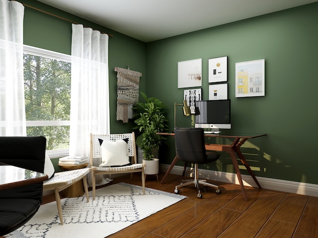 A green home office with a large window and white curtains on it with a computer desk in the right corner and a seating area right next to it.